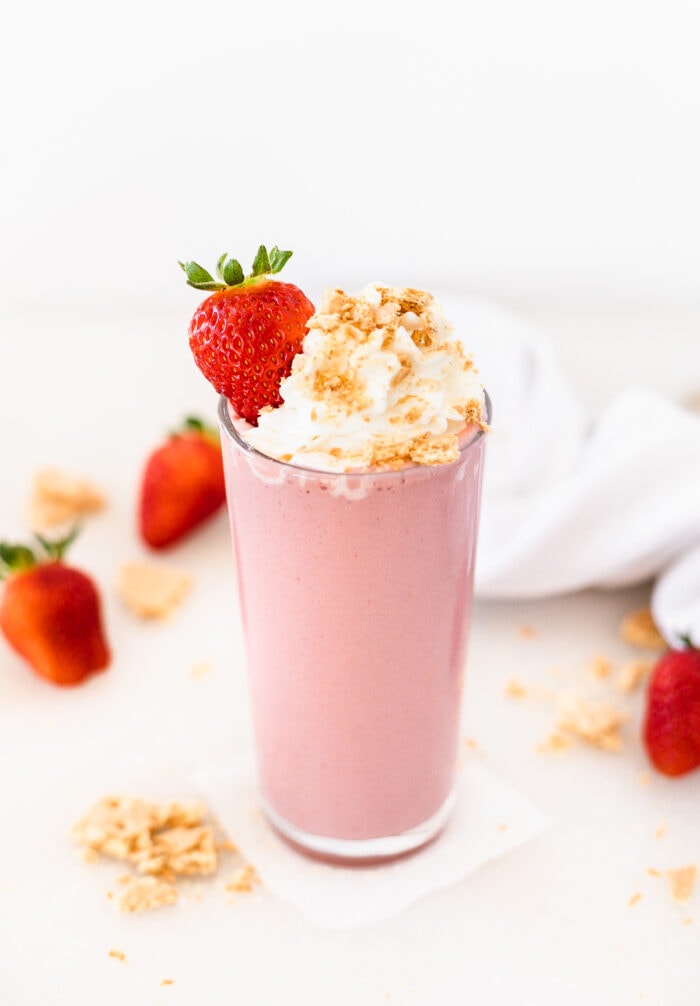 healthy strawberry cheesecake smoothie in a glass topped with whipped cream, graham cracker crumbs and a strawberry.