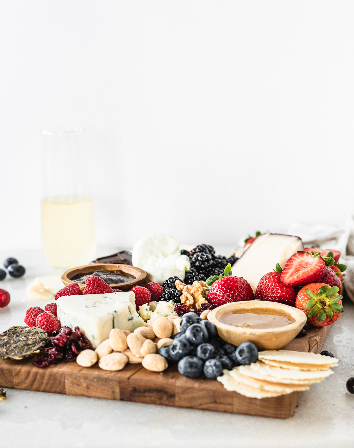 sweet and salty cheese board with berries, blue cheese, nuts, a bowl of honey and crackers with a glass of champagne in the background.