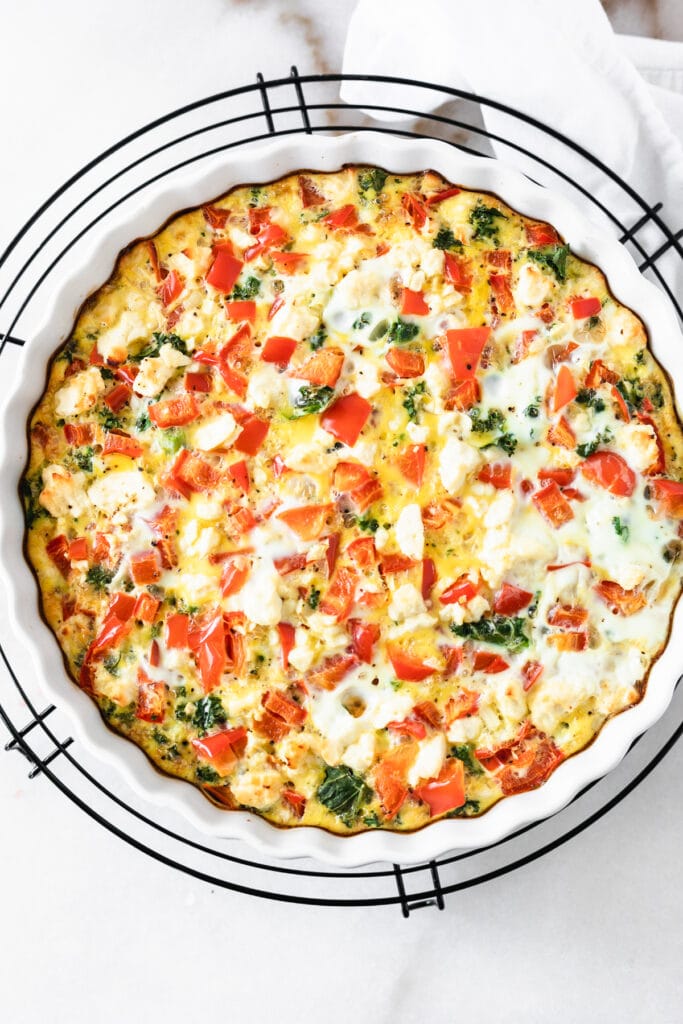 overhead view of red pepper kale and feta frittata in a round white baking dish on a black wire rack.
