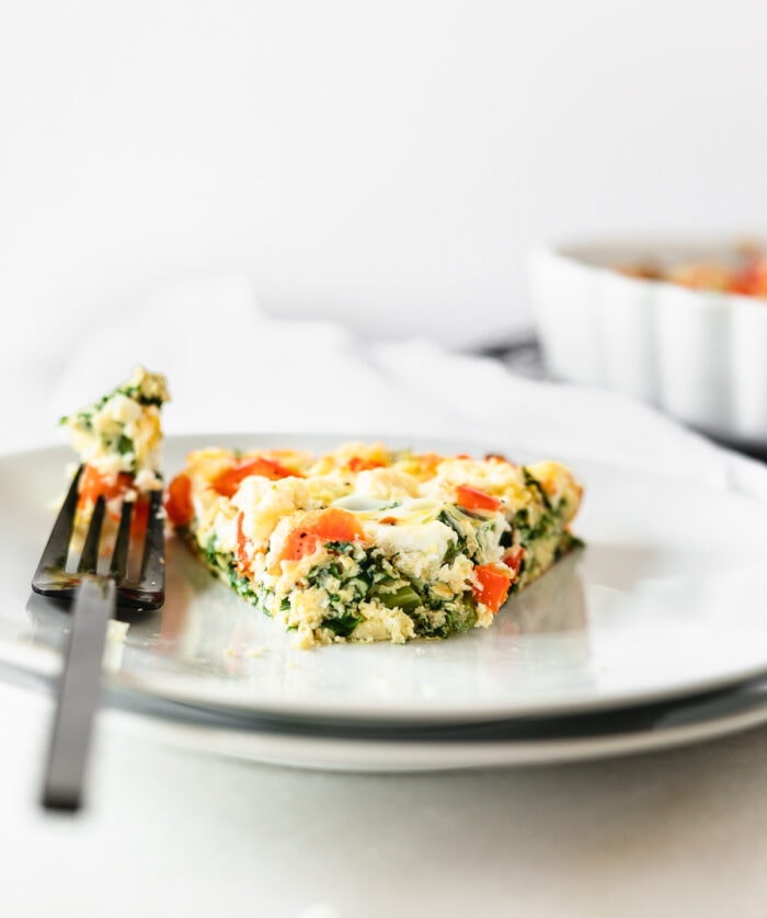 slice of kale red pepper feta frittata on a white plate with a bite taken out on a black fork sitting next to it.