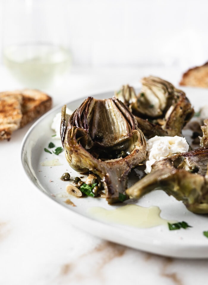 artichoke half with caper garlic white wine sauce on a plate with burrata and more artichokes.