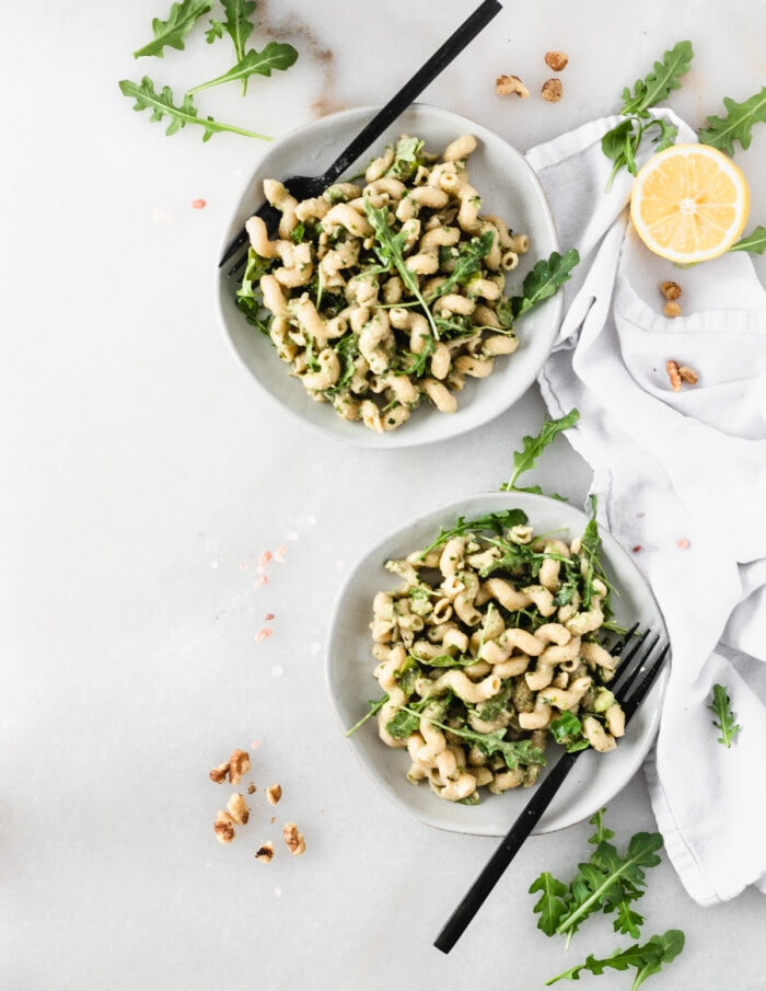 two plates of vegan arugula avocado pesto pasta with black forks in them surrounded by a white napkin, walnuts, arugula and lemon.