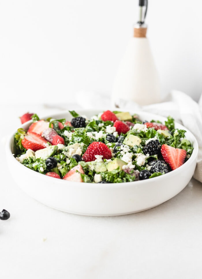 berry avocado kale salad in a white bowl with a bottle of olive oil in the background.