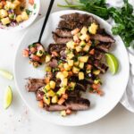 overhead view of sliced chipotle flank steak topped with pineapple salsa on a white plate with a spoon of salsa and lime wedges.