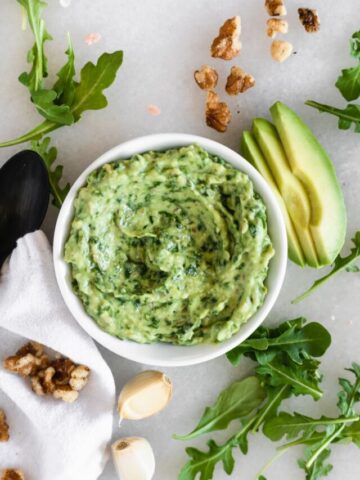 overhead view of vegan arugula avocado pesto in a white bowl surrounded by sliced avocado, arugula, walnuts, and garlic.