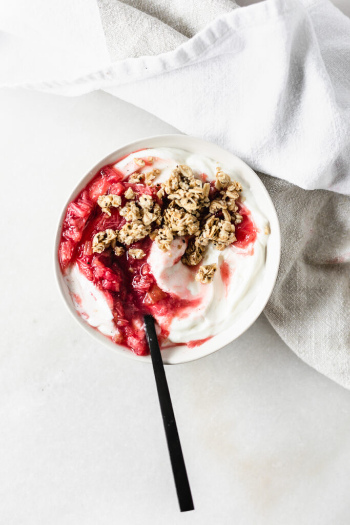 overhead view of a bowl of yogurt swirled with vanilla bean rhubarb compote and granola with a black spoon in it.