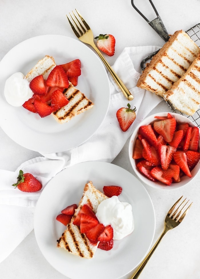 Strawberries and Cream Grilled Angel Food Cake on 2 white plates with a bowl of strawberries and gold forks.