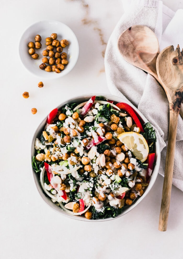 overhead view of moroccan roasted chickpea kale salad drizzled with tahini dressing with wooden spoons and a small bowl of roasted chickpeas next to it.