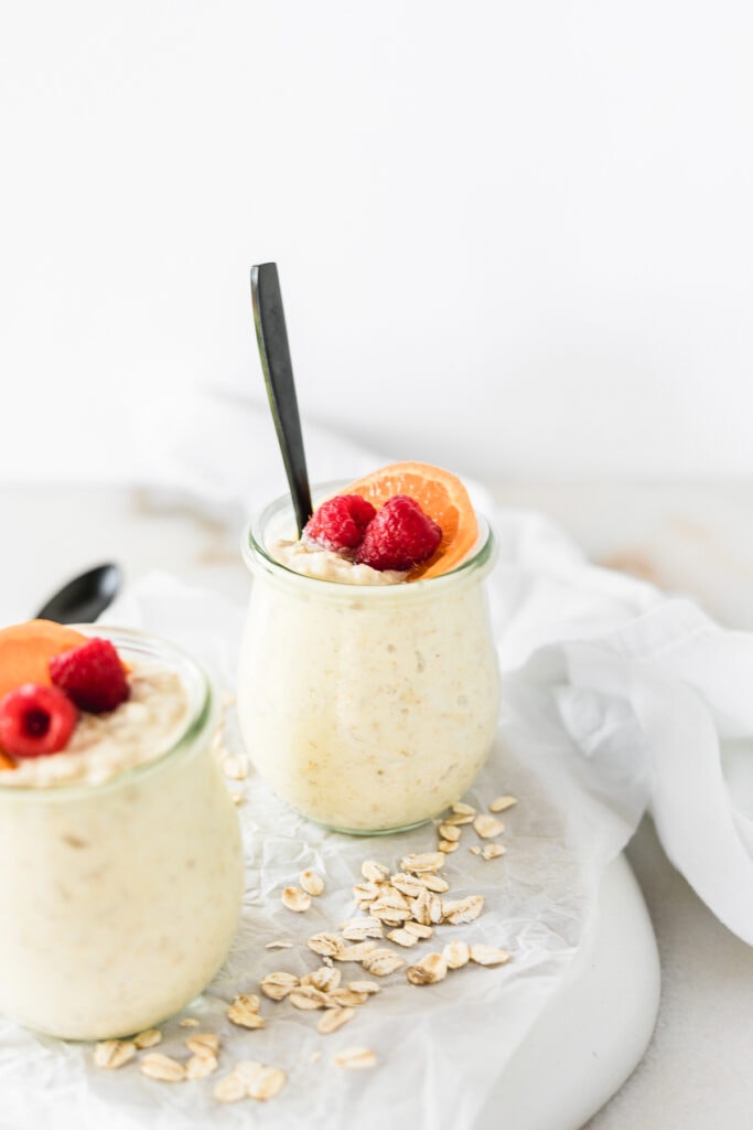 Orange Creamsicle Overnight Oats topped with an orange slice and raspberries in a glass jar with a black spoon in it.