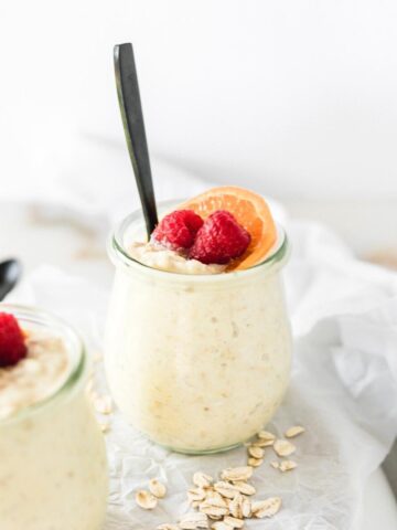 Orange Creamsicle Overnight Oats topped with an orange slice and raspberries in a glass jar with a black spoon in it.