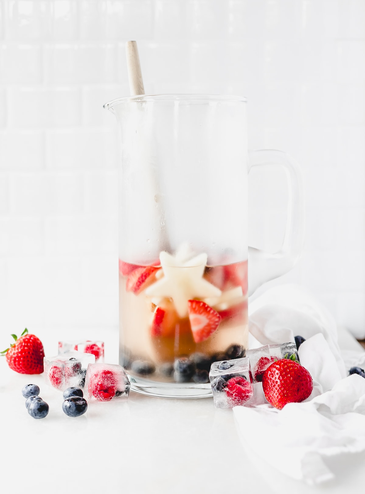 pitcher of red white and blue sangria with a wood spoon in it surrounded by berries.