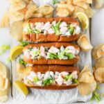 overhead view of 3 new england style shrimp rolls surrounded by potato chips and lemon wedges.