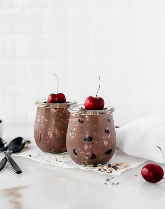 two glass jars with black forest overnight oats topped with cherries on a white background.