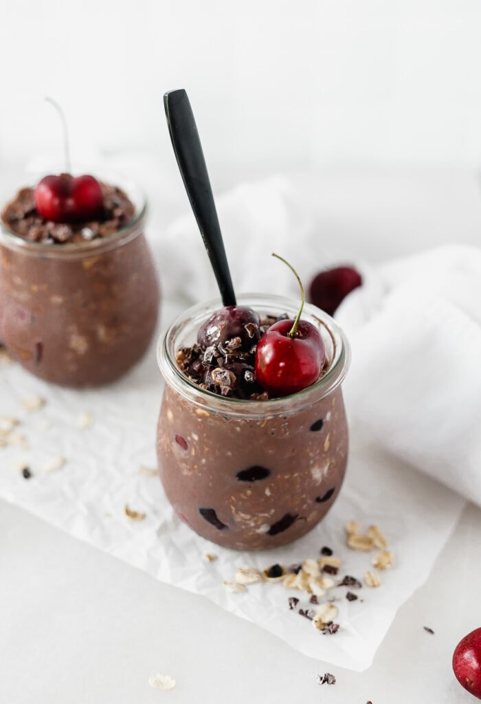 black forest overnight oats in a glass jar with a cherry and cacao nibs on top and a black spoon in it.
