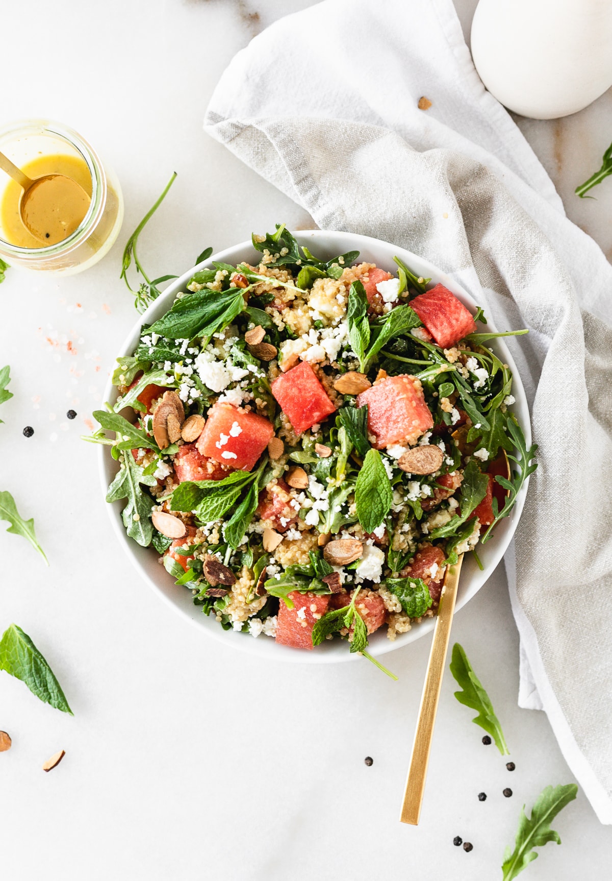overhead view of watermelon arugula salad with quinoa, feta, almonds and mint in a white bowl with a gold spoon in it.