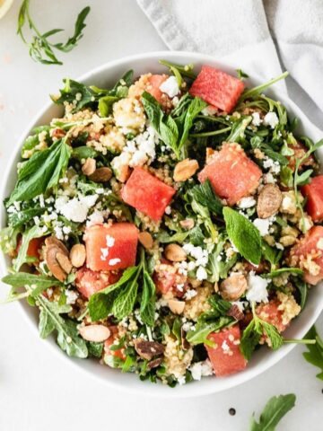 overhead view of watermelon arugula salad with feta, quinoa, almonds and mint in a white bowl.