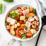 overhead view of corn peach and tomato salad in a white bowl with a black spoon beside it.