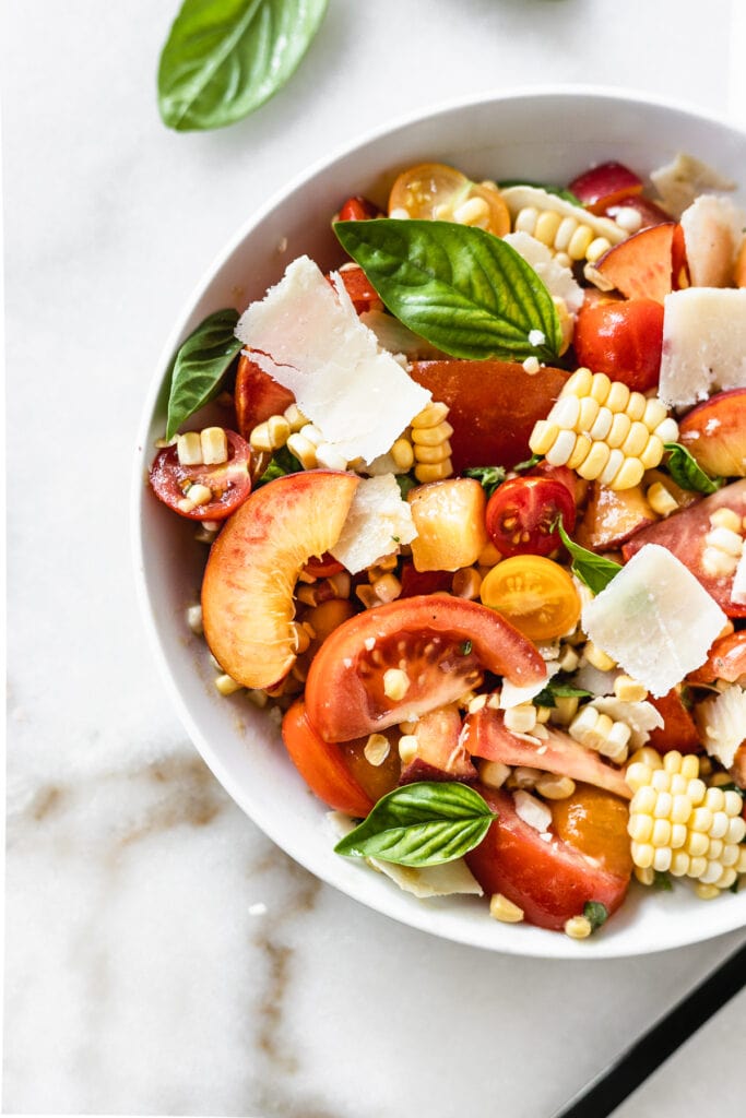 closeup overhead view of corn peach and tomato basil salad with shaved parmesan in a white bowl.