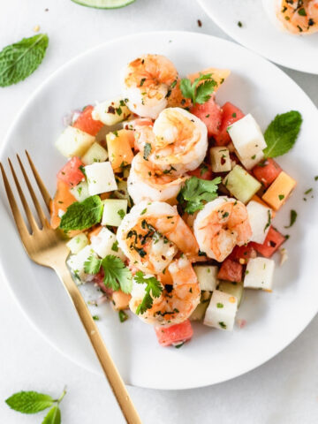 overhead view of grilled shrimp and jicama melon salad on a white plate with a gold fork.