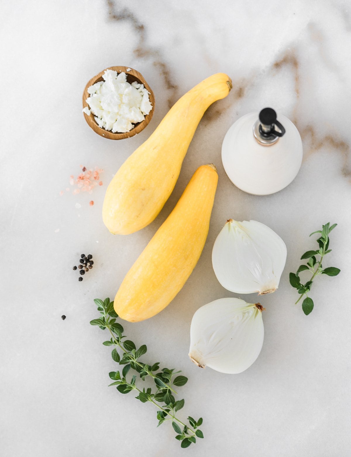 overhead view of two yellow squash, a halved onion, a bottle of olive oil, oregano and a bowl of goat cheese crumbles on a white background. 