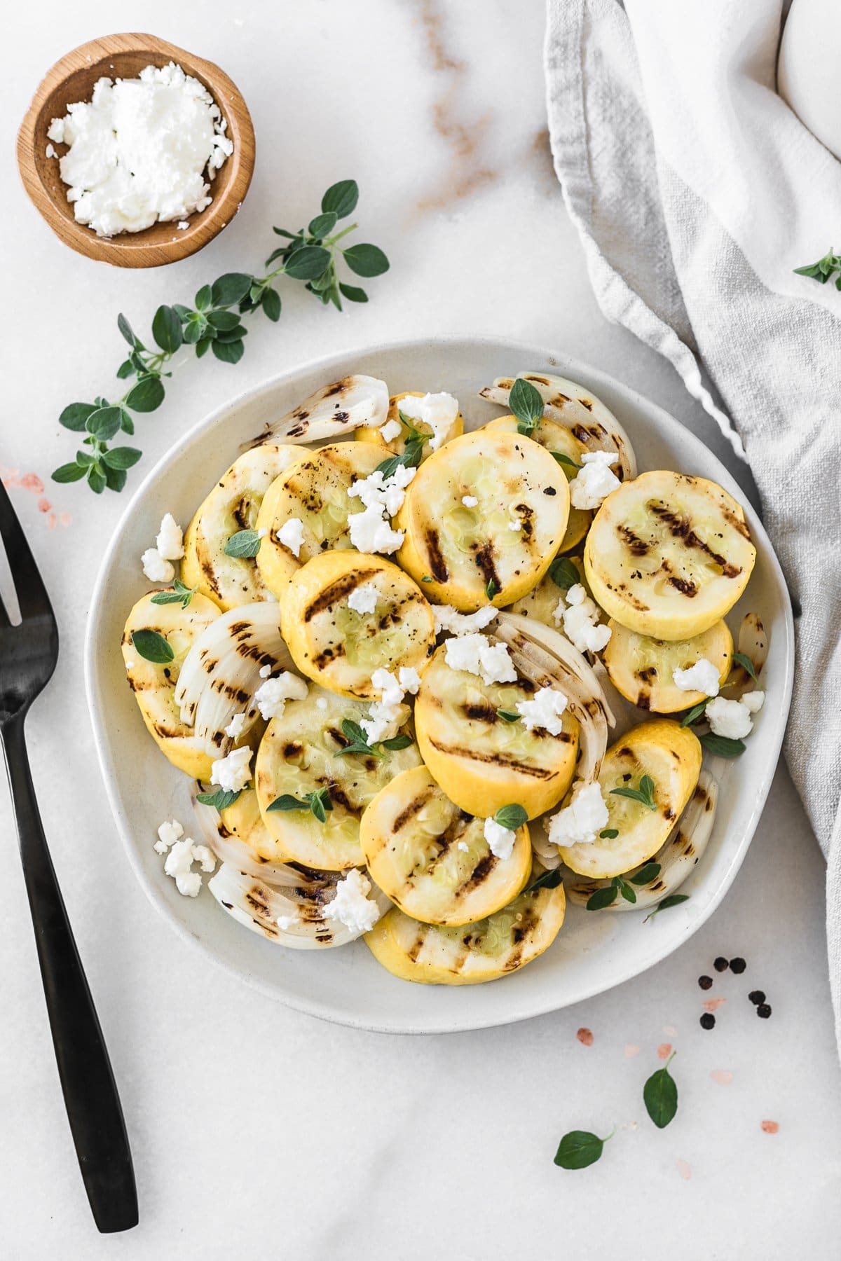 overhead view of grilled yellow squash and onions on a plate topped with goat cheese and oregano with a black fork, bowl of goat cheese and oregano sprigs next to it.