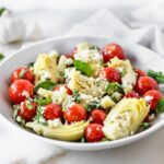 marinated artichoke salad with tomatoes herbs and feta in a white bowl.
