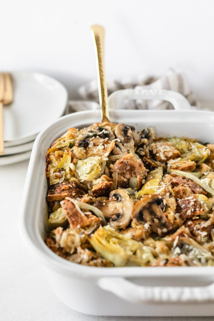 artichoke mushroom strata in a white baking dish with a gold serving spoon in it.