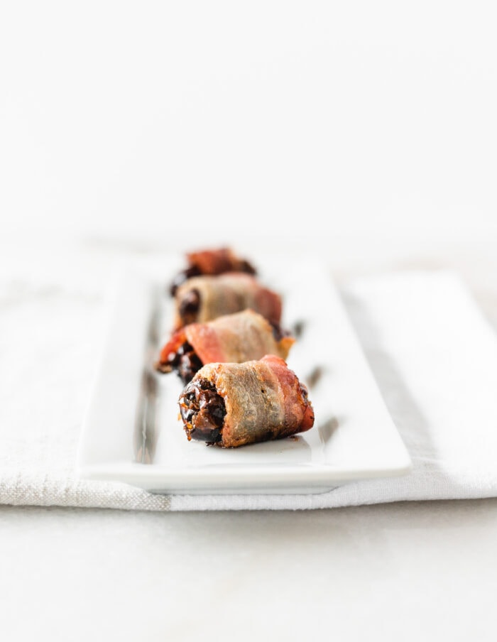bacon wrapped dates in a line on a white tray.