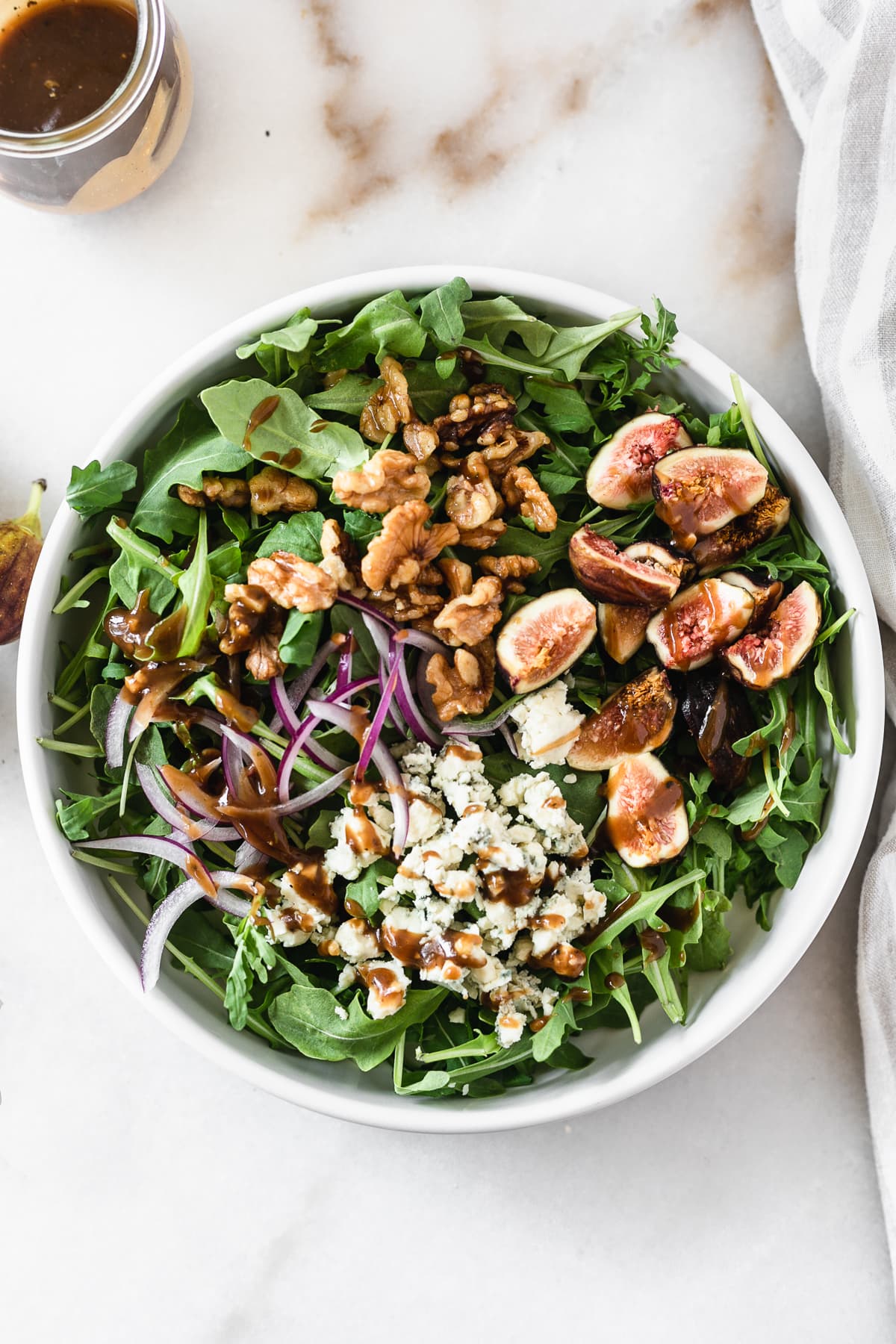 overhead view of arugula in a white bowl topped with quartered figs, candied walnuts, red onion, blue cheese and balsamic vinaigrette.