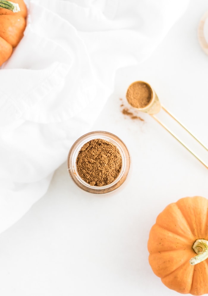 homemade pumpkin pie spice in a small glass jar with a teaspoon and pumpkins beside it.