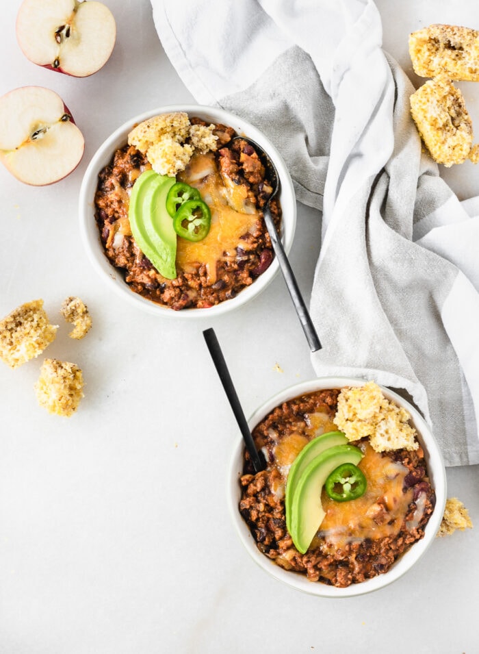 overhead view of two bowls of chipotle apple turkey chili topped with cheese, avocado, jalapeño and cornbread with black spoons in them.