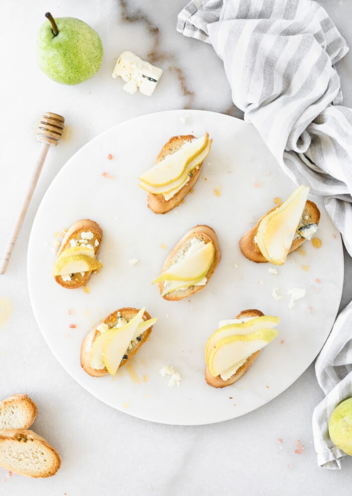 pear blue cheese crostini on a white serving board surrounded by bread slices, a honey dipper, pears, blue cheese, and a striped linen.