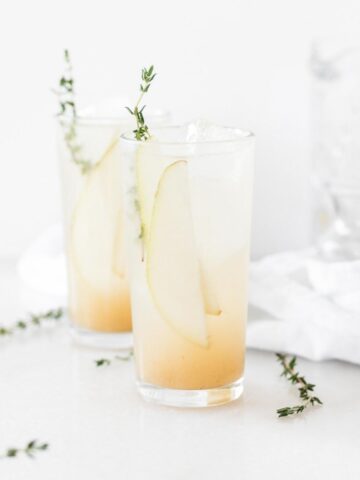 two pear collins cocktails in highball glasses garnished with pear slices and thyme.