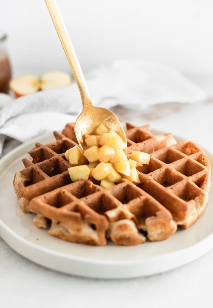 gold spoon placing caramel apple topping onto a waffle on a white plate.