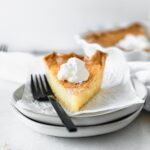 slice of buttermilk pie topped with whipped cream next to a black fork on a stack of plates.