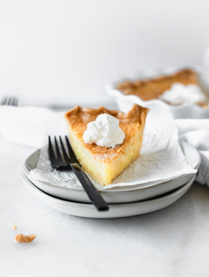 slice of buttermilk pie topped with whipped cream next to a black fork on a stack of plates.