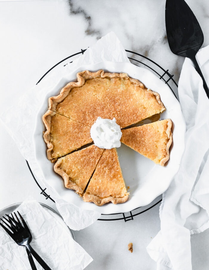 overhead view of a sliced buttermilk pie topped with whipped cream in a white pie dish surrounded by a pie server, black forks, and a white linen.