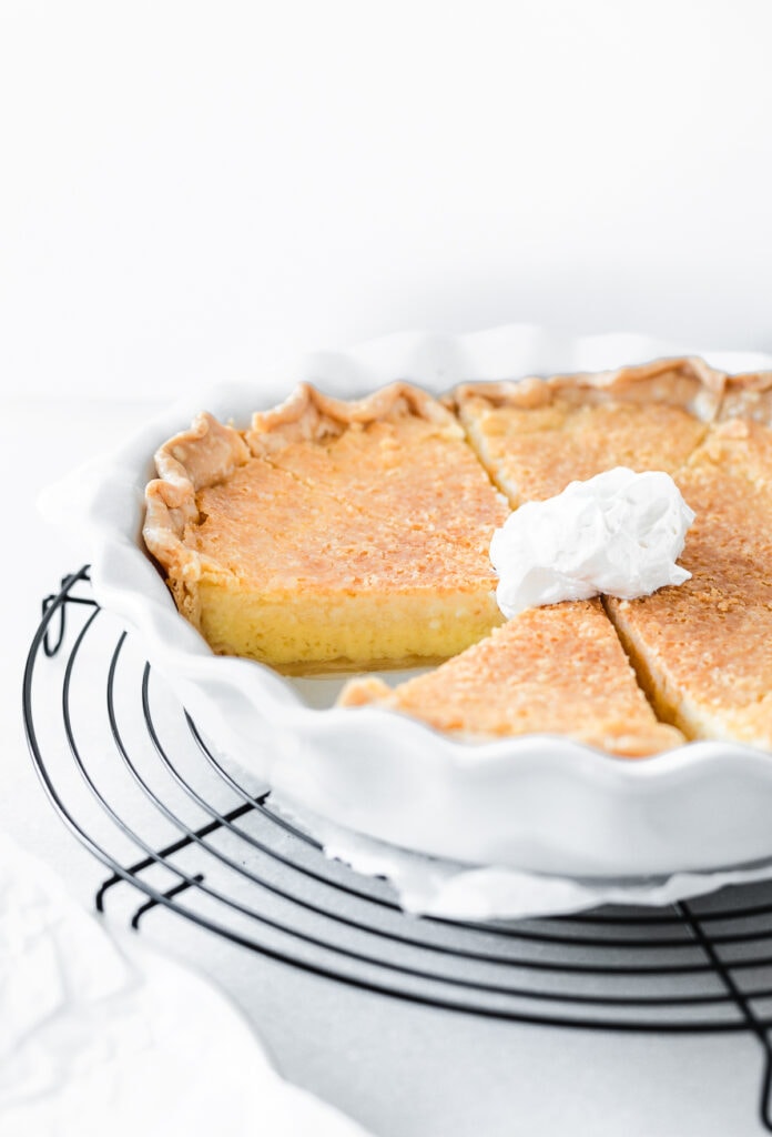 sliced southern buttermilk pie in a white pie dish with whipped cream on top on a black cooling rack.