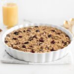 cranberry orange baked oatmeal in a round white baking dish with a glass of orange juice in the background.