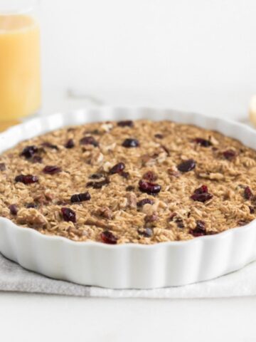 cranberry orange baked oatmeal in a round white baking dish with a glass of orange juice in the background.