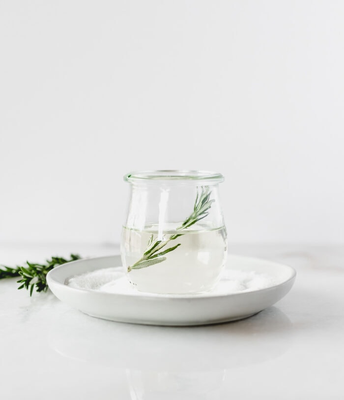 rosemary simple syrup in a glass jar with a sprig of rosemary in it on top of a grey plate.