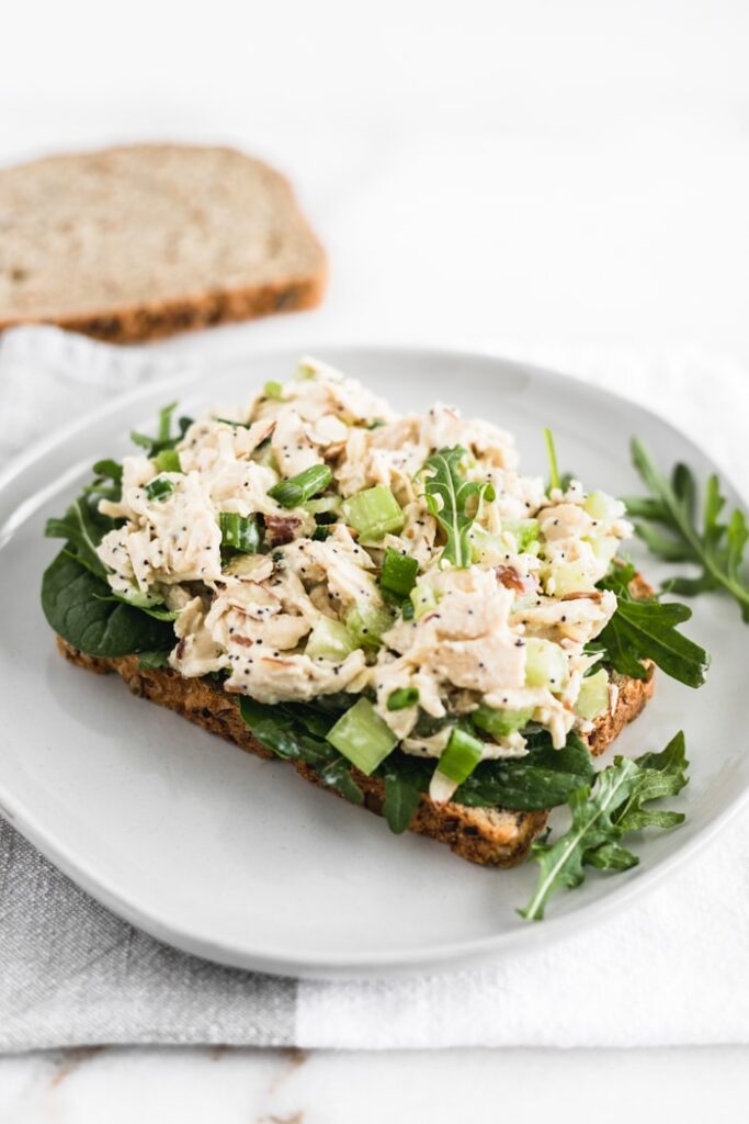 no mayo orange poppy seed chicken salad on a piece of bread with greens on a white plate.