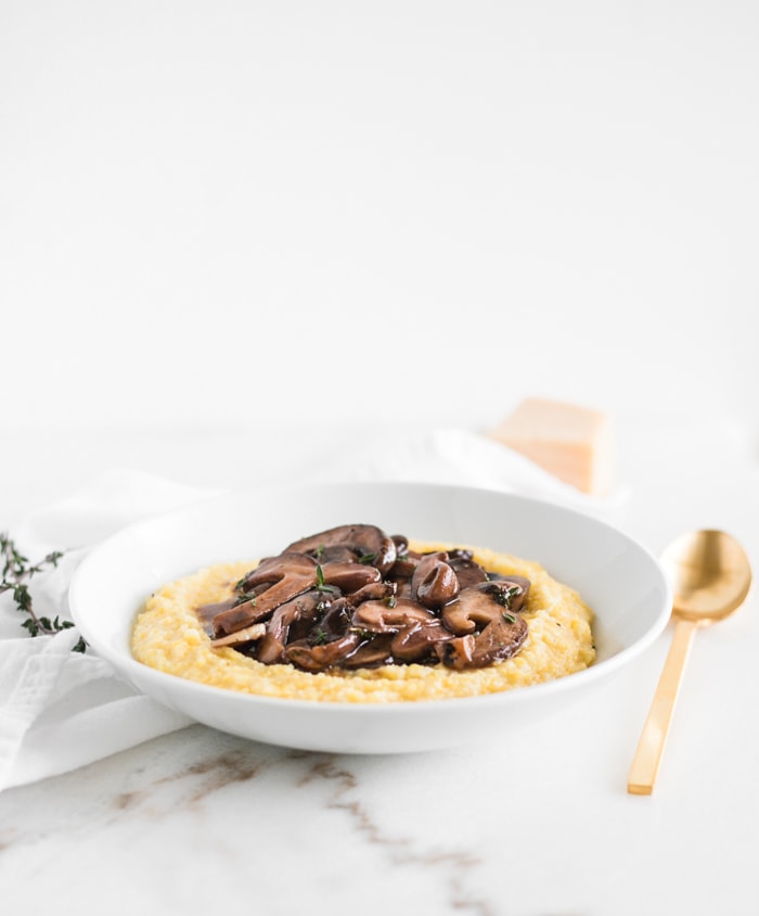 white bowl with creamy parmesan polenta topped with red wine mushrooms surrounded by a gold spoon, white napkin, thyme, and a chunk of parmesan.