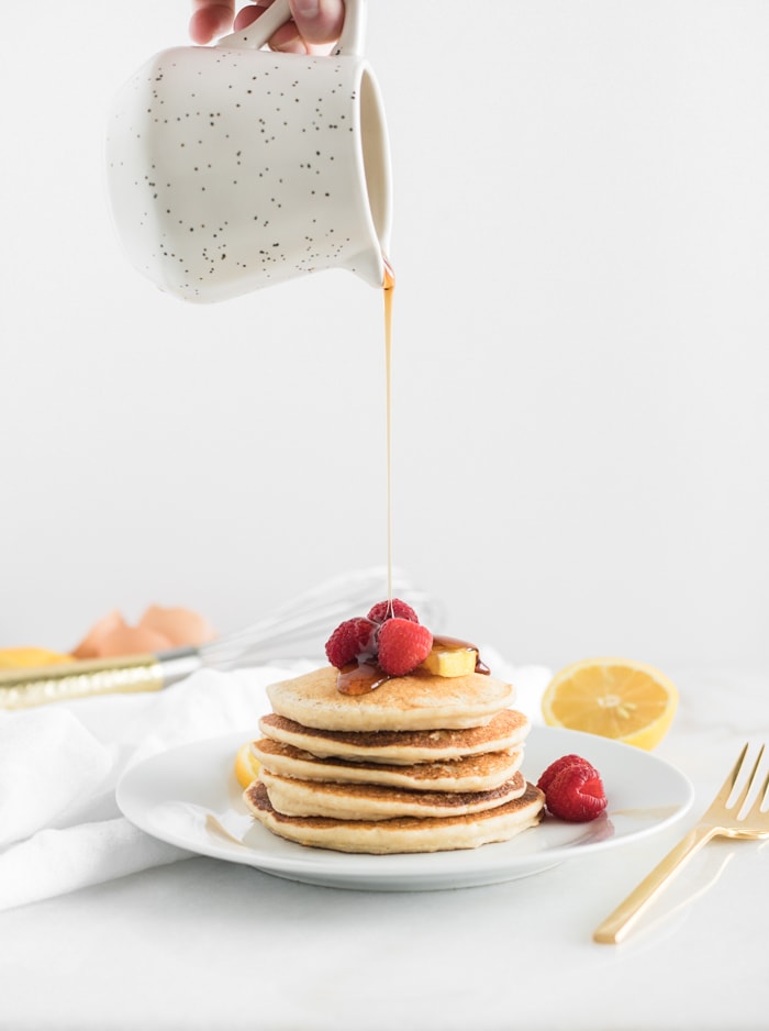 syrup being poured on a stack of healthy lemon ricotta pancakes with raspberries on top.