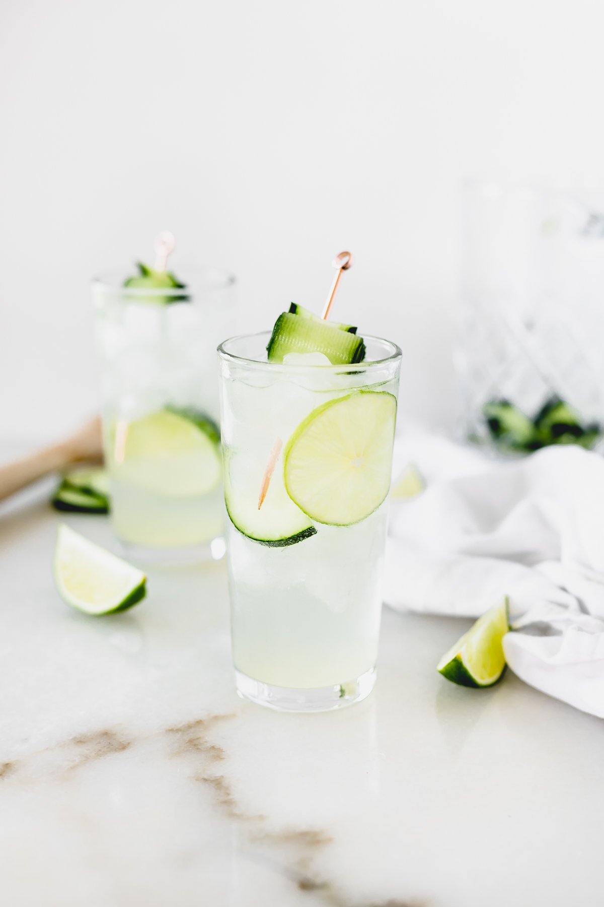 white linen cocktail in a glass garnished with lime and cucumber surrounded by lime wedges.