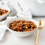 homemade baked beans in a white bowl with a gold spoon beside it.