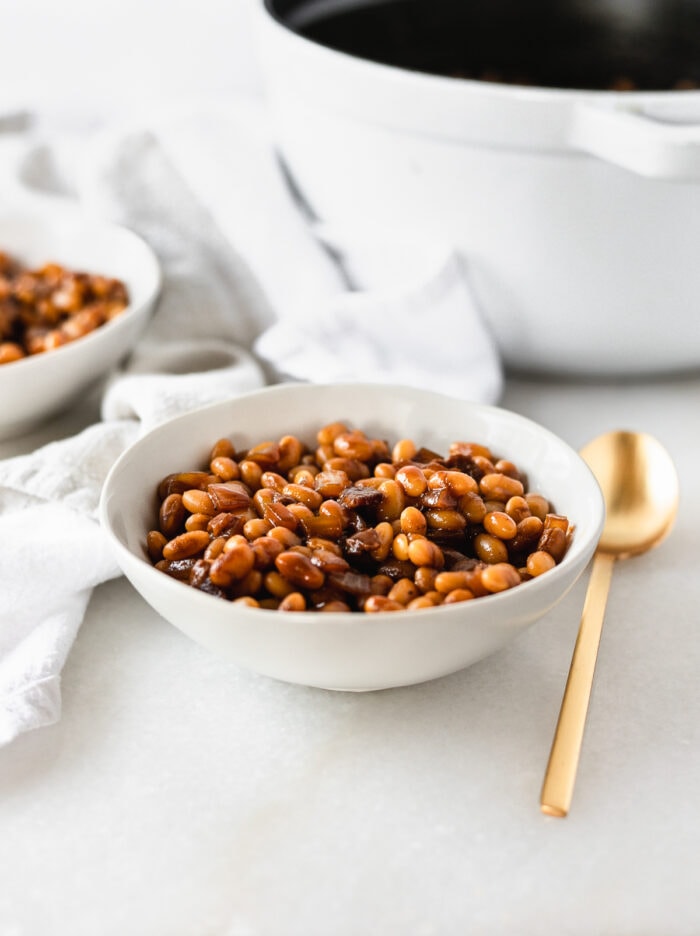 homemade baked beans in a white bowl with a gold spoon beside it.