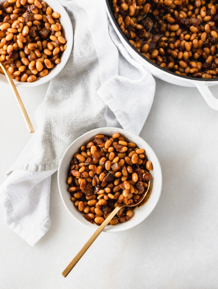 overhead view of homemade baked beans in two white bowls with gold forks on a white background.