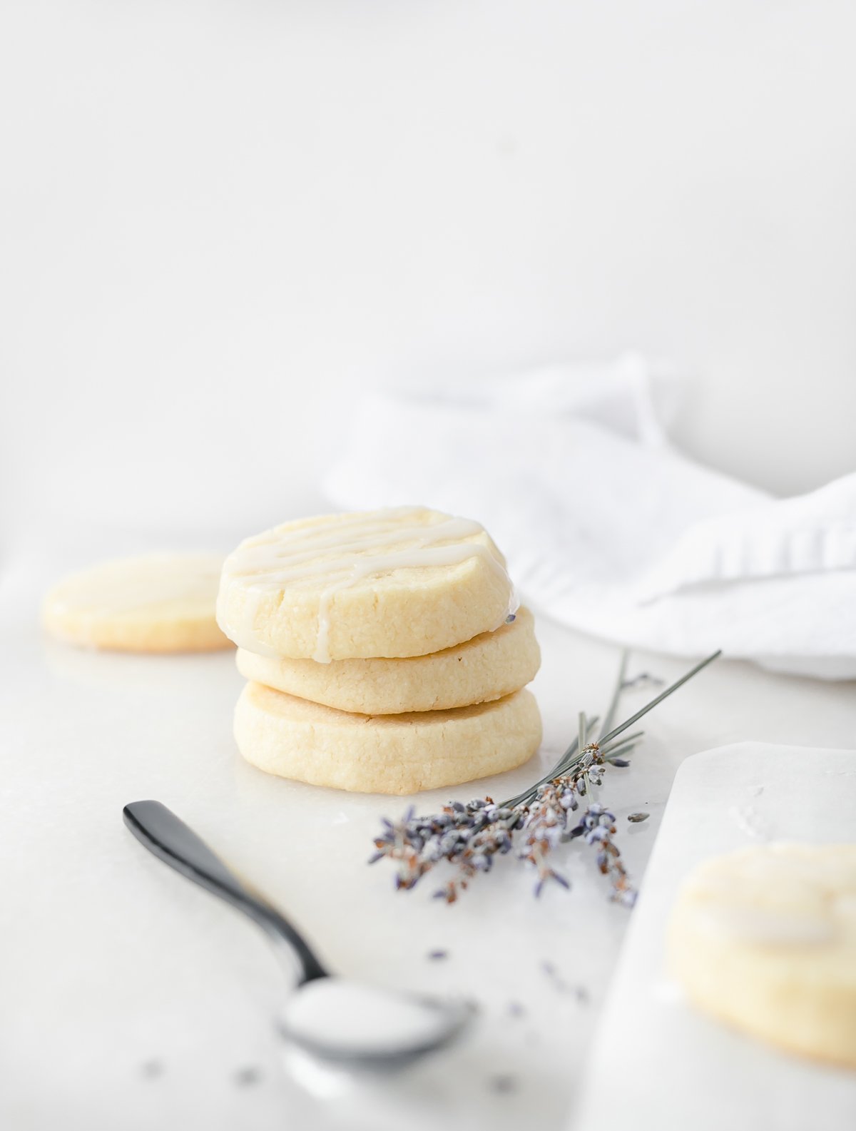 three stacked lavender shortbread cookies with dried lavender sprigs beside them and a spoonful of glaze in the foreground.