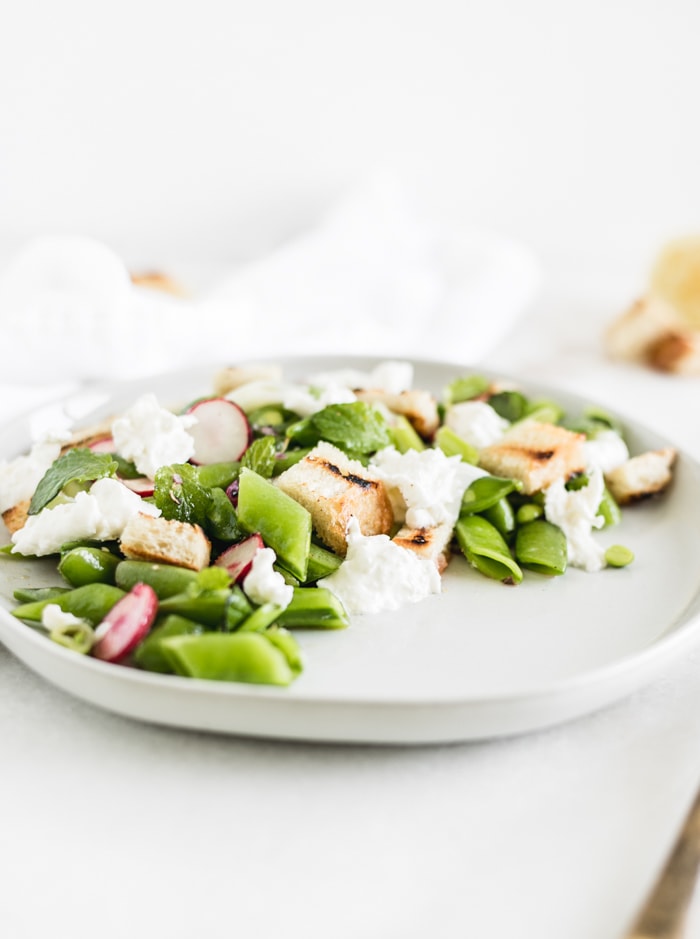 snap pea salad with burrata, sourdough croutons, and lemon vinaigrette on a white plate.